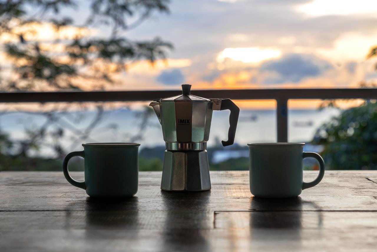 A moka pot flanked by two enamel mugs on a weathered wooden table, silhouetted against a sunrise over the sea